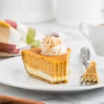 A slice of decadent pumpkin cheesecake topped with whipped cream and a sprinkle of cinnamon sits on a white plate. A fork rests next to the slice, with a bite of the cheesecake on it. The background features a colorful cloth and a white mug, enhancing the cozy autumnal vibe.