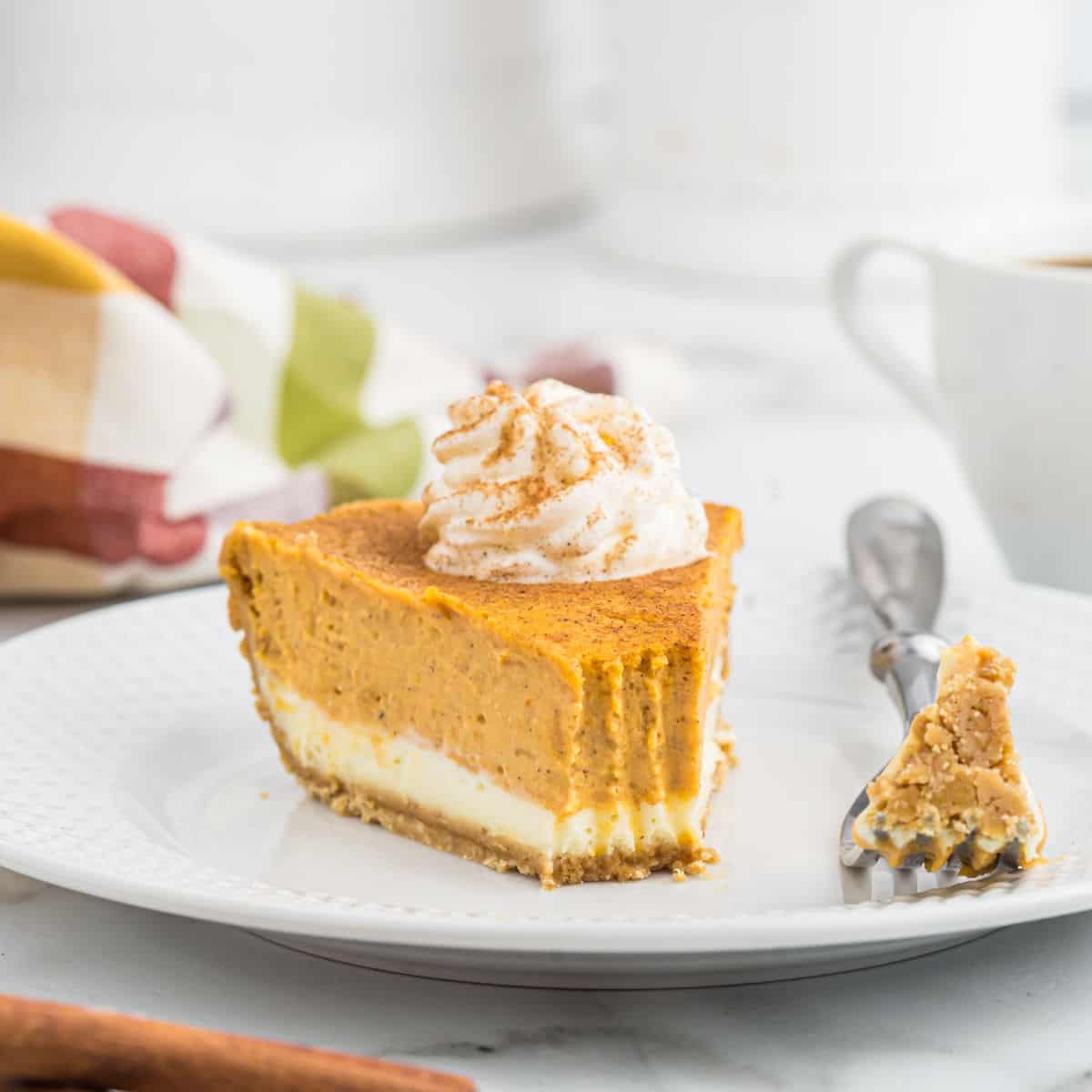 A slice of decadent pumpkin cheesecake topped with whipped cream and a sprinkle of cinnamon sits on a white plate. A fork rests next to the slice, with a bite of the cheesecake on it. The background features a colorful cloth and a white mug, enhancing the cozy autumnal vibe.