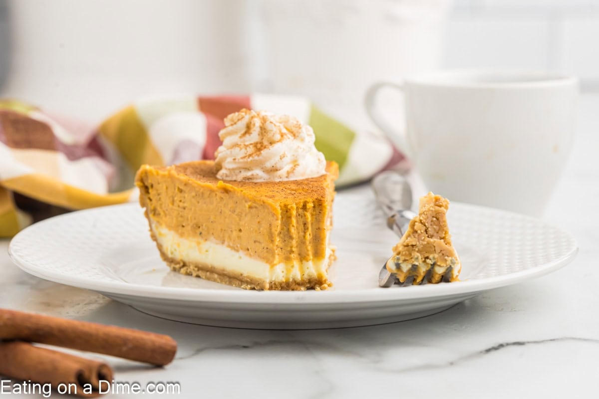 A slice of layered pumpkin cheesecake topped with whipped cream on a white plate with a fork holding a piece. The background showcases a colorful striped cloth and a white mug, with cinnamon sticks nearby. Text at the bottom reads "Eating on a Dime.com.