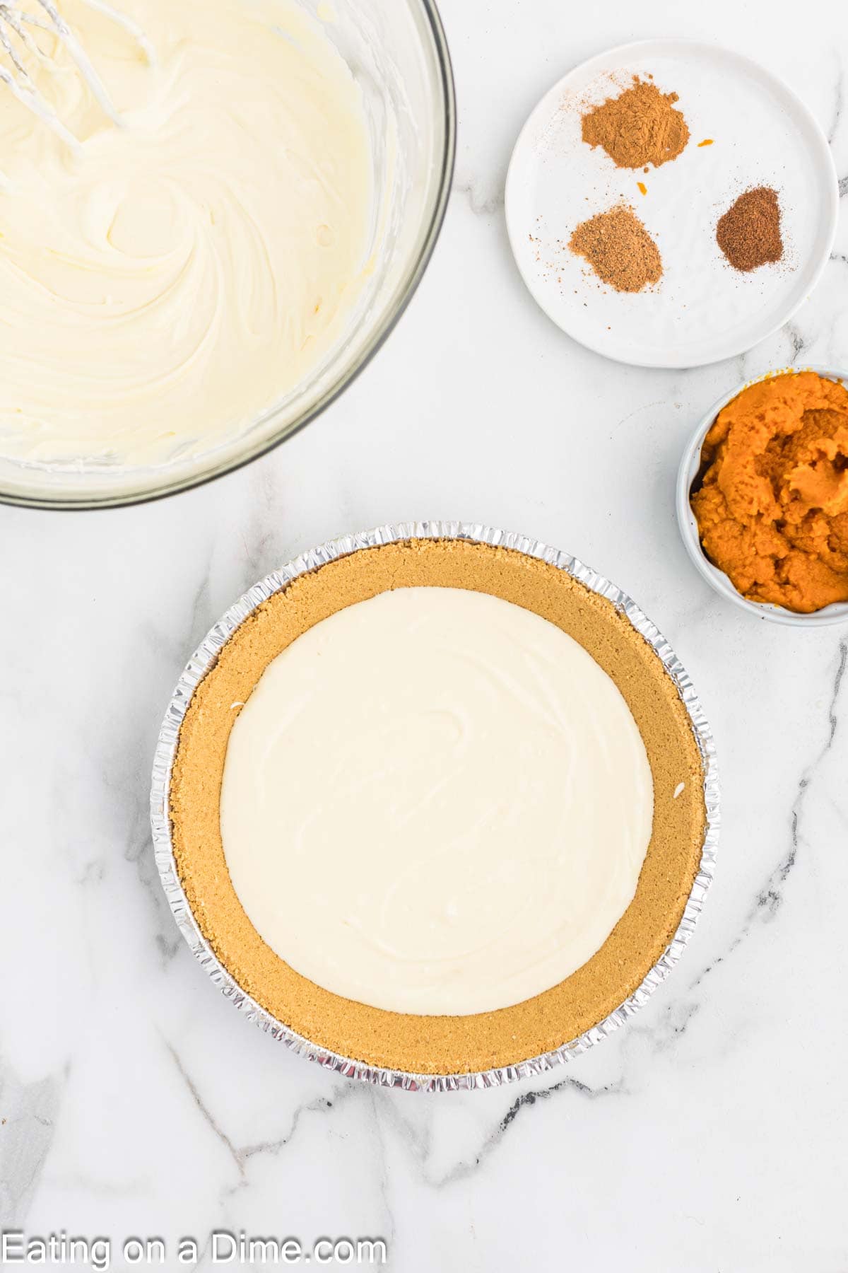 A graham cracker pie crust filled with a creamy mixture rests on a white marble surface. Nearby, bowls hold whipped cream mixture, pumpkin puree, and three types of spices. The ingredients are being prepared for a delectable pumpkin cheesecake recipe.