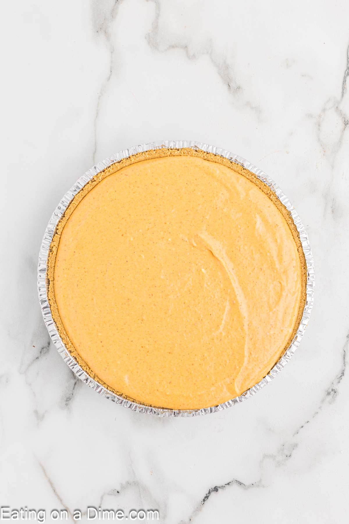 A round pumpkin pie in a silver foil pie tin sits on a white marble surface. The pie has a smooth, plain orange filling that hints at the creamy decadence of pumpkin cheesecake and features a thin, slightly visible crust.