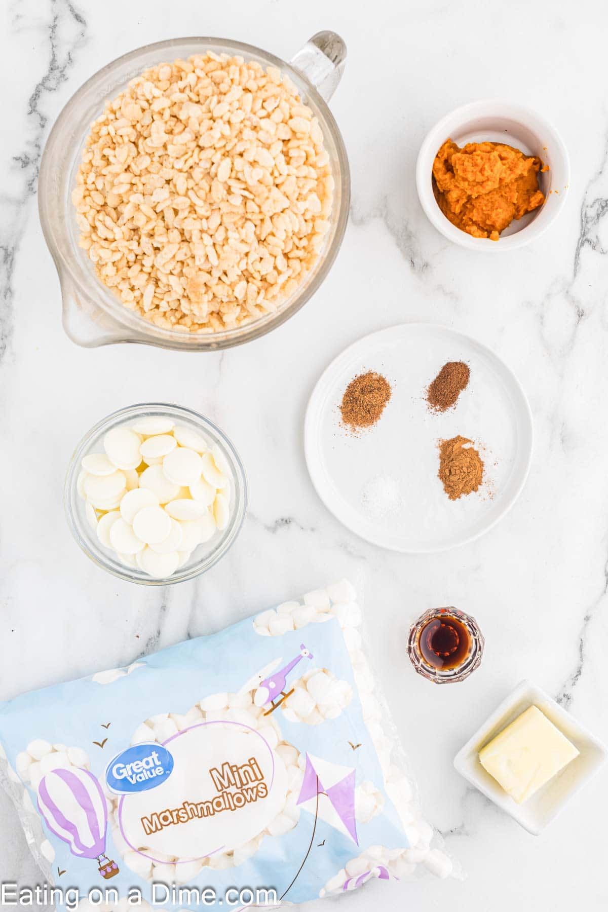 A marble surface displays ingredients for pumpkin rice krispie treats: a mixing bowl of rice cereal, a small white bowl of white chocolate chips, a package of mini marshmallows, a white plate with spices, a bowl of pumpkin puree, a dish with vanilla extract, and a pat of butter.