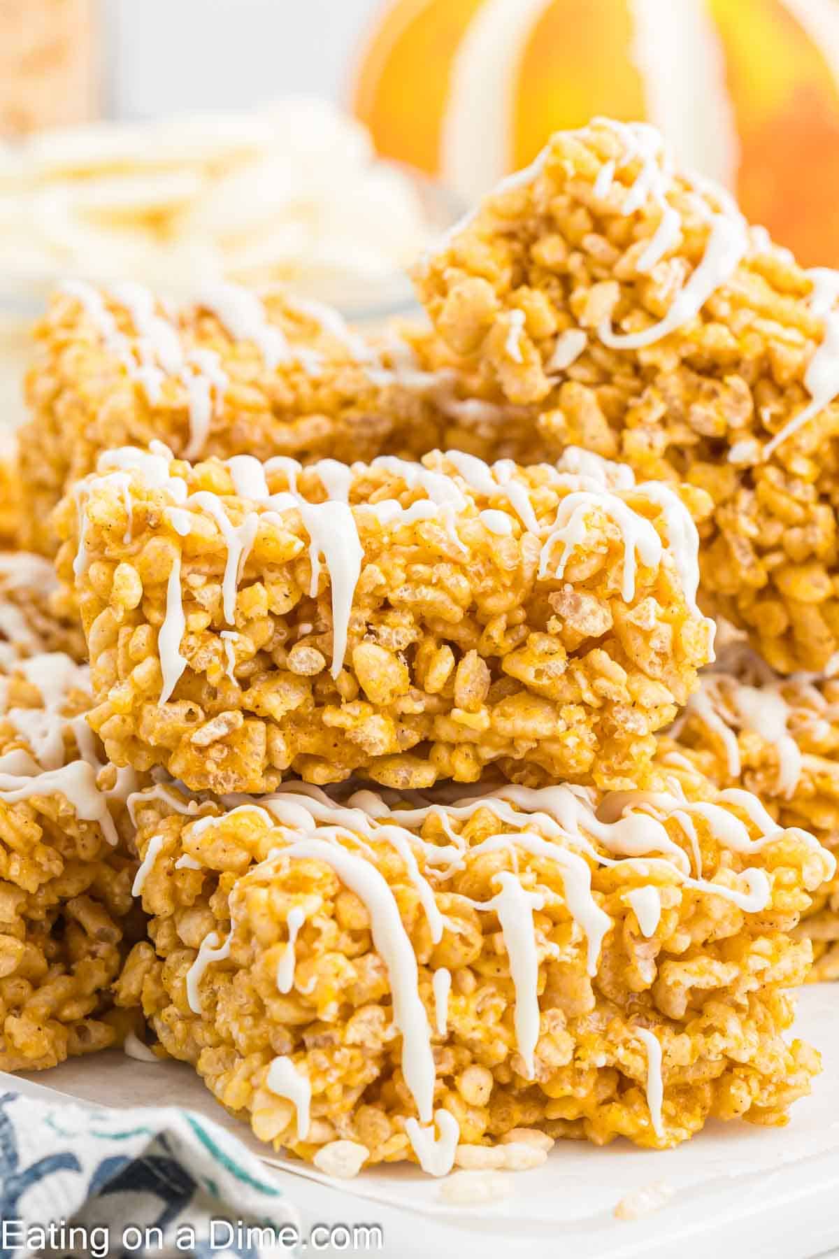 A close-up of pumpkin rice krispie treats stacked on a plate, drizzled with white icing. The treats are golden brown and textured with puffed rice, and a blurred background shows hints of oranges and other food items.