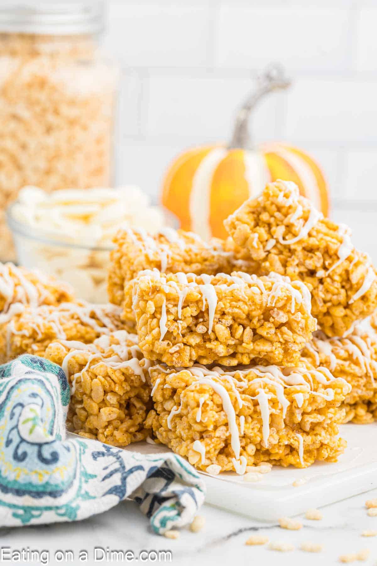 A stack of pumpkin rice krispie treats drizzled with white icing sits on a white table. A glass jar of rice cereal and a small pumpkin are decoratively placed in the background. A colorful cloth napkin lies next to the treats. A branding watermark reads "Eating on a Dime.com.