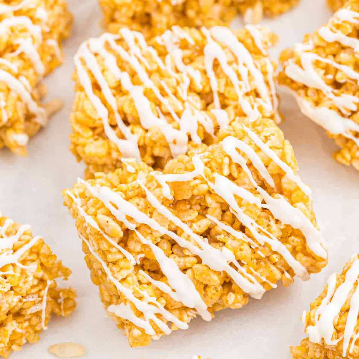 Close-up of a batch of golden pumpkin rice krispie treats, drizzled with white icing. The treats are arranged in a slightly scattered manner on a light surface, showcasing their crispy texture and sweet glaze.