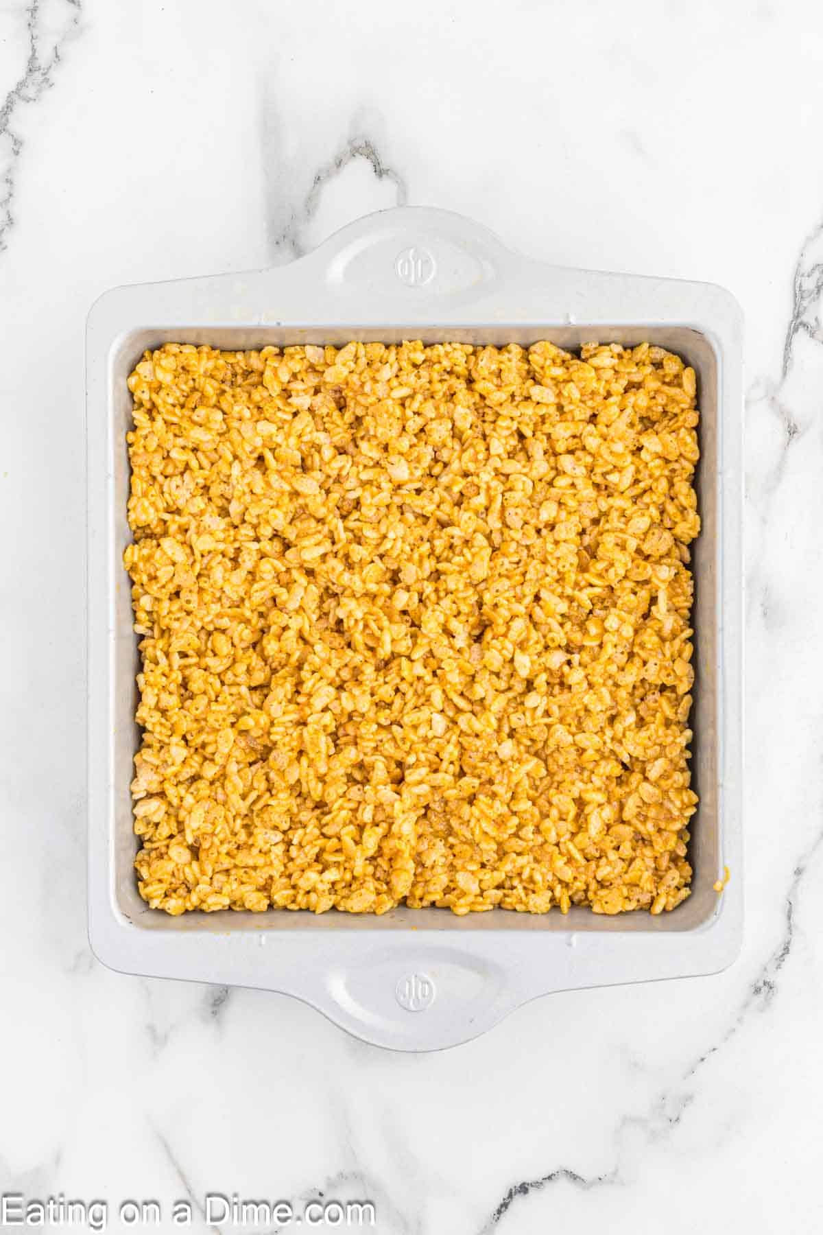 A square baking dish filled with golden brown pumpkin Rice Krispie treats rests on a marble countertop. The crispy, marshmallow-coated cereal squares, infused with a hint of pumpkin spice, fill the entire dish, ready to be cut and served.