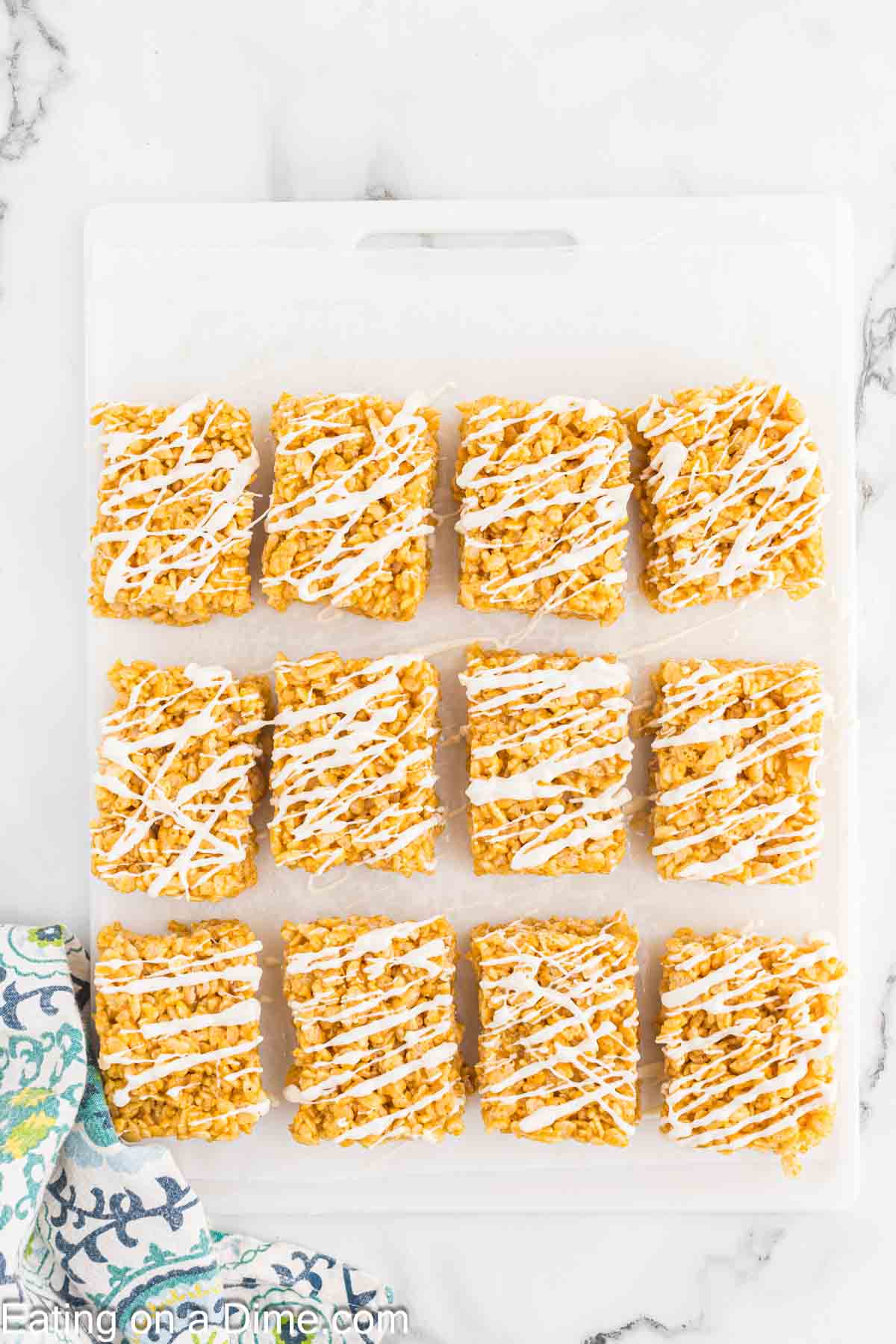 Twelve pumpkin rice krispie treats drizzled with white icing are neatly arranged in three rows on a white surface. A colorful floral-patterned cloth is partially visible in the bottom left corner.