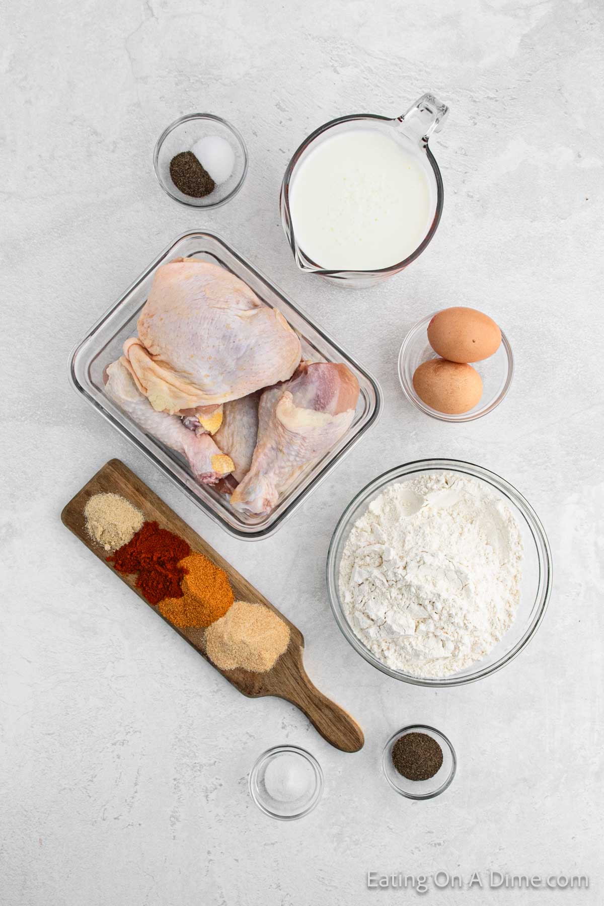 Overhead view of raw chicken pieces in a glass container, a bowl of flour, two eggs, a measuring cup with liquid, and several small bowls of spices and seasonings arranged on a light-colored surface. A wooden board holds more spices and seasoning blends—perfect for preparing air fryer fried chicken.