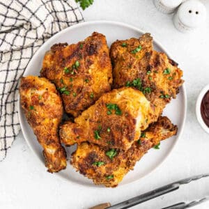 A plate of golden-brown air fryer fried chicken pieces garnished with chopped parsley, placed on a white surface. A checkered napkin and salt and pepper shakers are visible in the background, alongside a small bowl of dipping sauce and a pair of tongs.