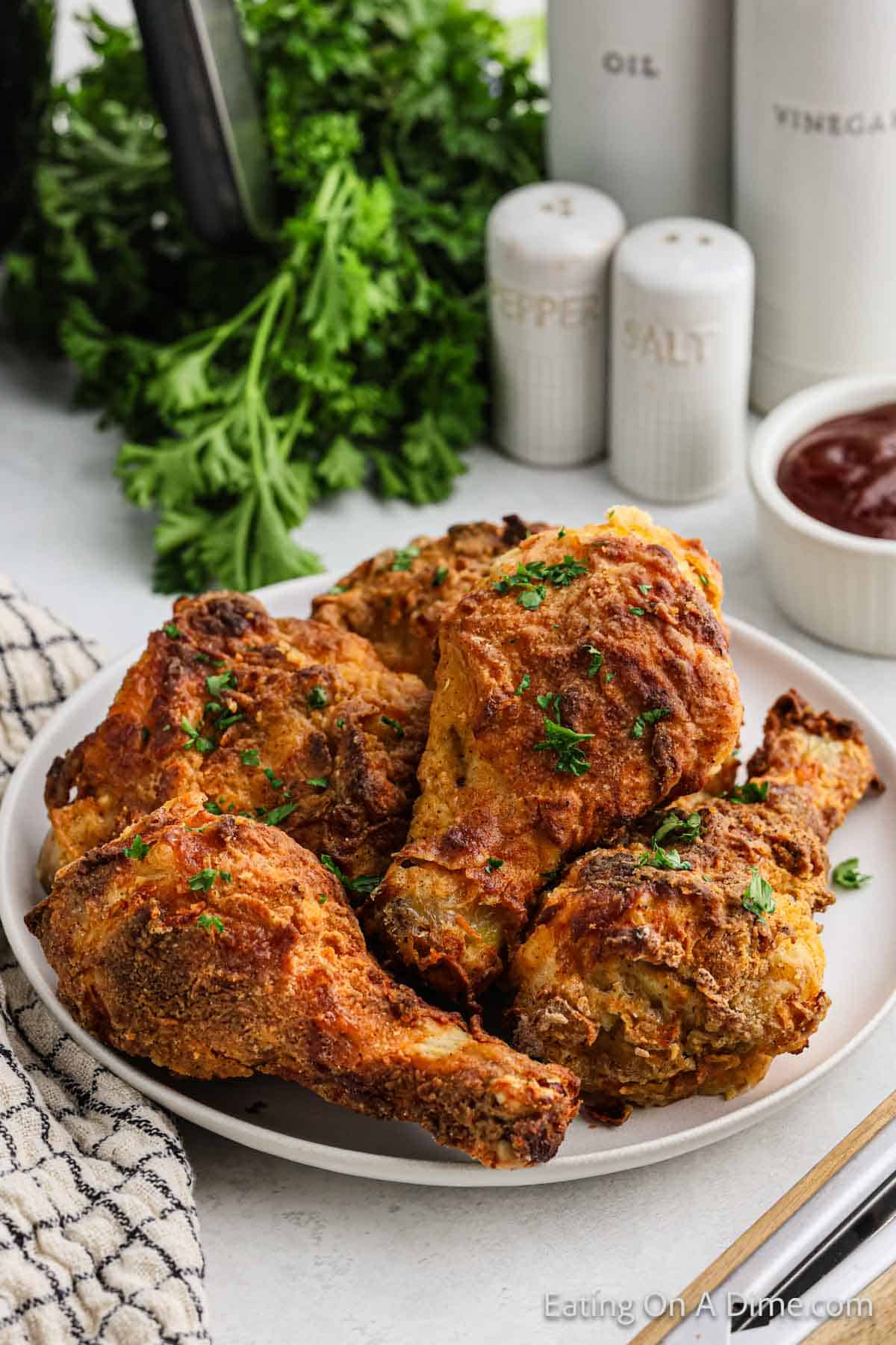 A plate filled with crispy air fryer fried chicken drumsticks garnished with herbs. Behind the plate are a napkin, a bunch of fresh parsley, and condiment containers labeled oil, vinegar, pepper, and salt. A small bowl filled with sauce is also visible.