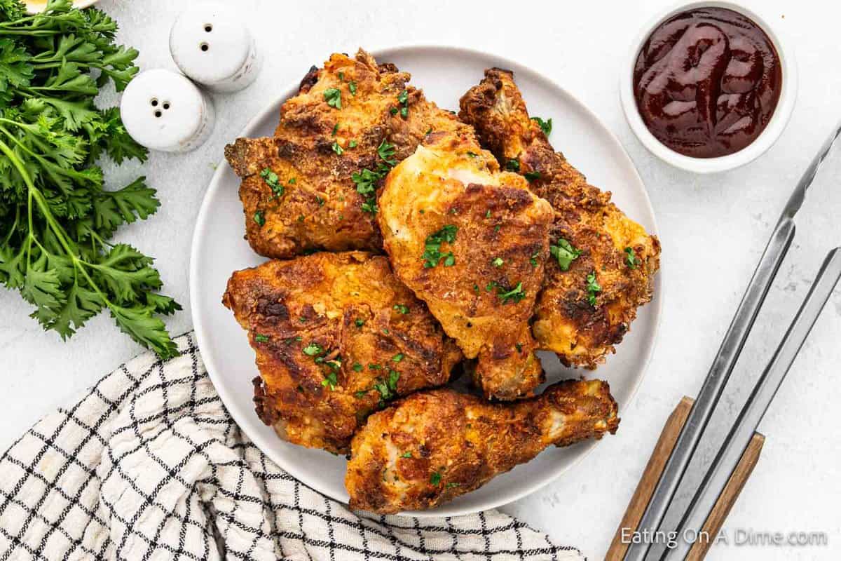 A plate of air fryer fried chicken pieces garnished with chopped parsley, accompanied by a small bowl of barbecue sauce. Fresh parsley, salt and pepper shakers, and a checkered cloth napkin are in the background along with a pair of metal tongs.