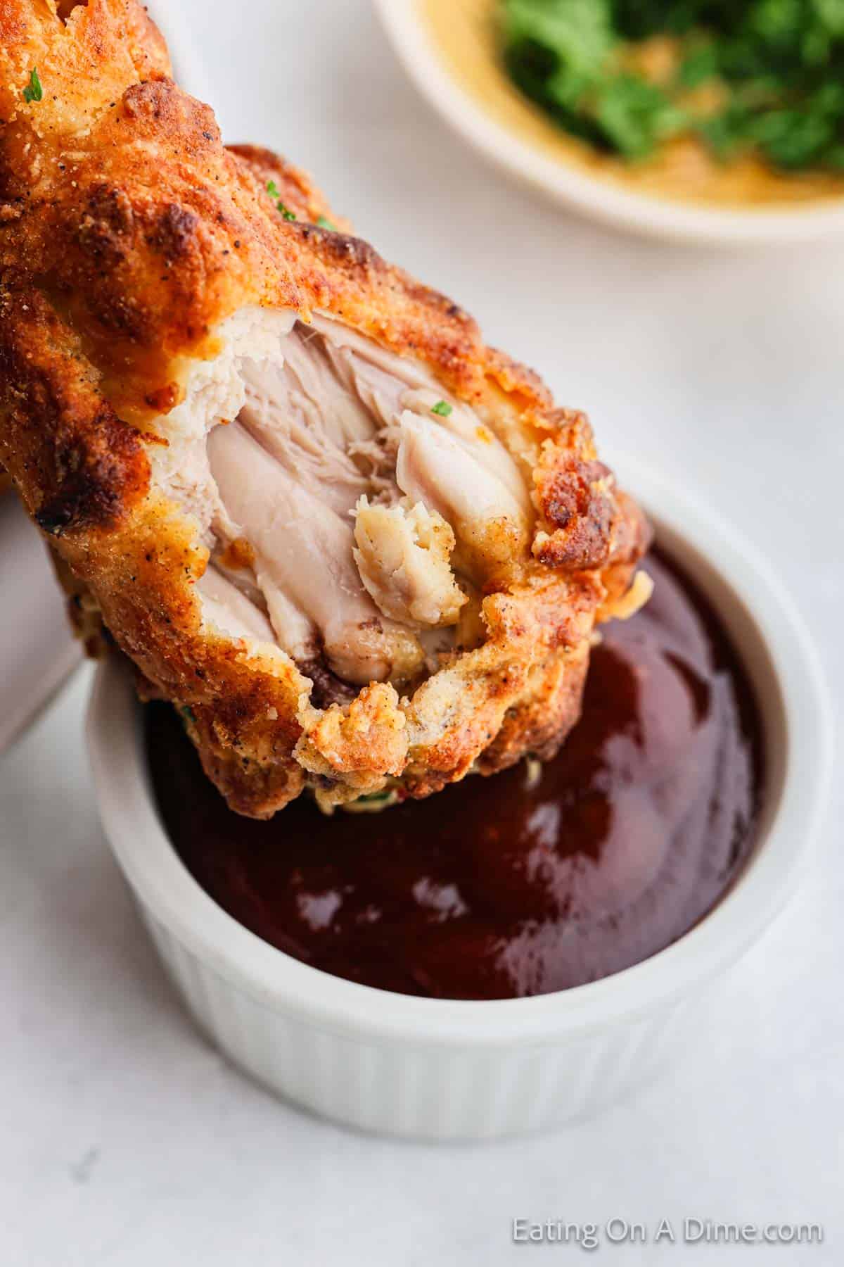 A crispy air fryer fried chicken drumstick, partially bitten into, is being dipped into a cup of dark barbecue sauce. In the background, there is a bowl with green herbs. The setting is on a light surface, possibly a kitchen counter or table.