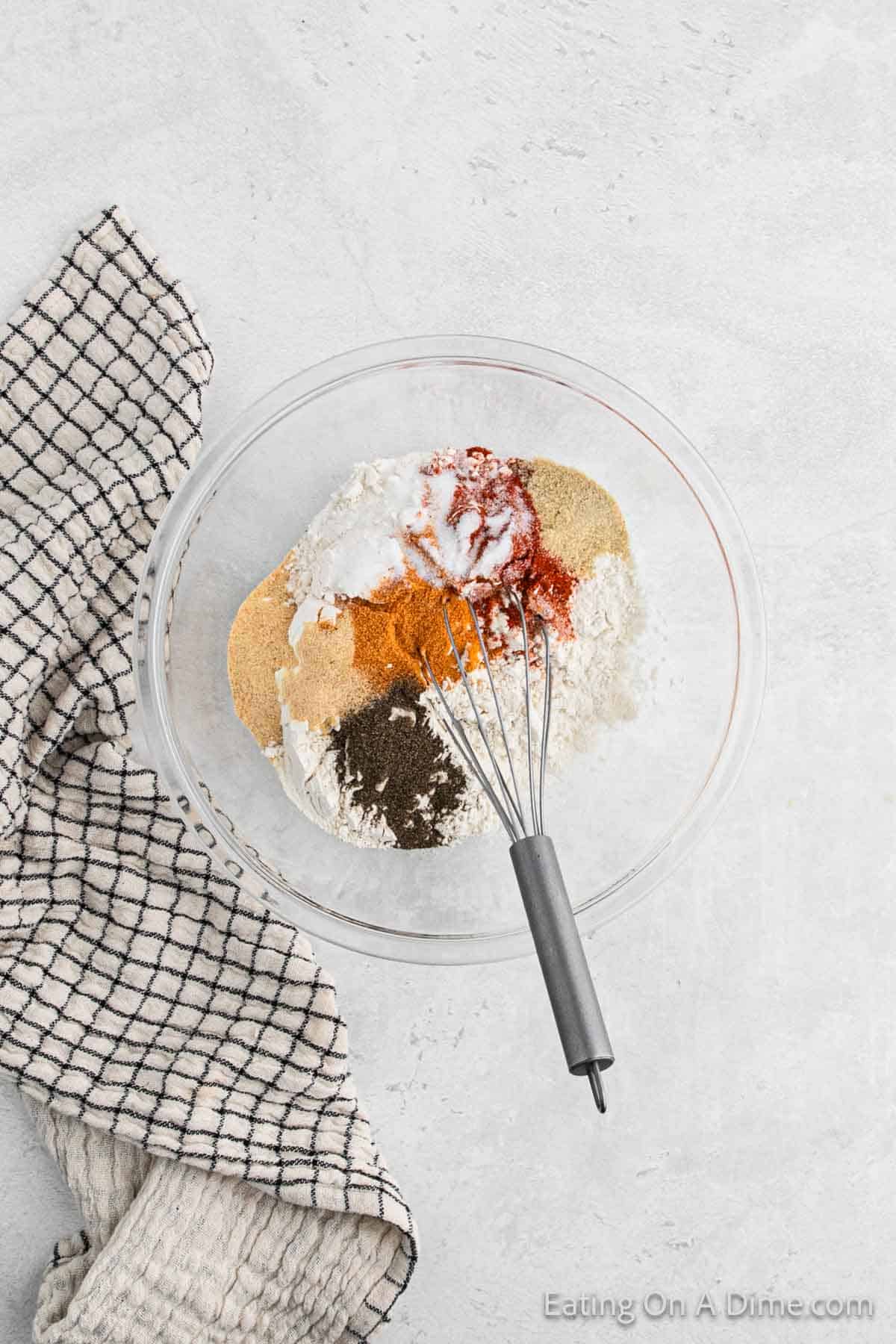 A large glass bowl filled with various spices and flour sits on a light gray surface, perfect for prepping air fryer fried chicken. A metal whisk rests against the side of the bowl, and a black-and-white checkered cloth is placed to the left.