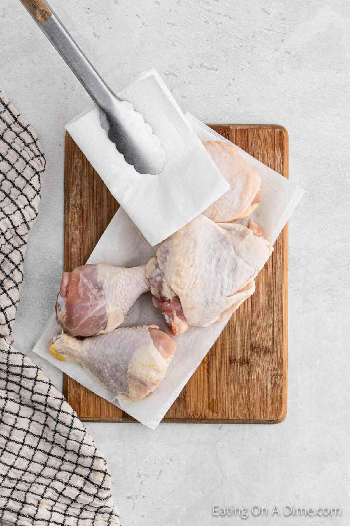 A pair of metal tongs with a white napkin beside raw chicken thighs on a wooden cutting board, ready to become delicious air fryer fried chicken. A black and white checkered cloth is partially visible on the left side, and the background is a light-colored surface.