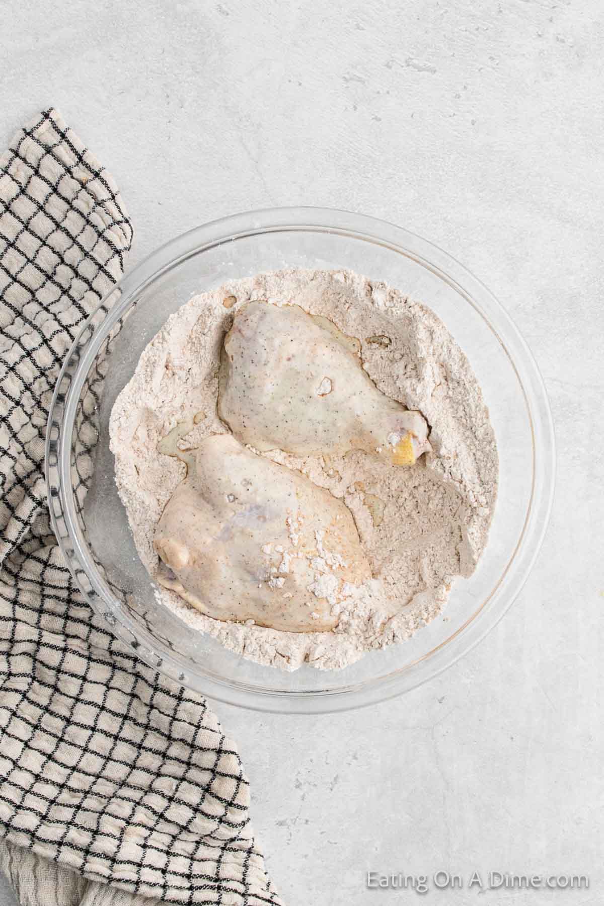 Two raw chicken drumsticks, ready to transform into delicious air fryer fried chicken, sit coated in flour and seasoning in a clear glass bowl on a light gray surface. A black-and-white checkered kitchen towel is partially visible to the left of the bowl.