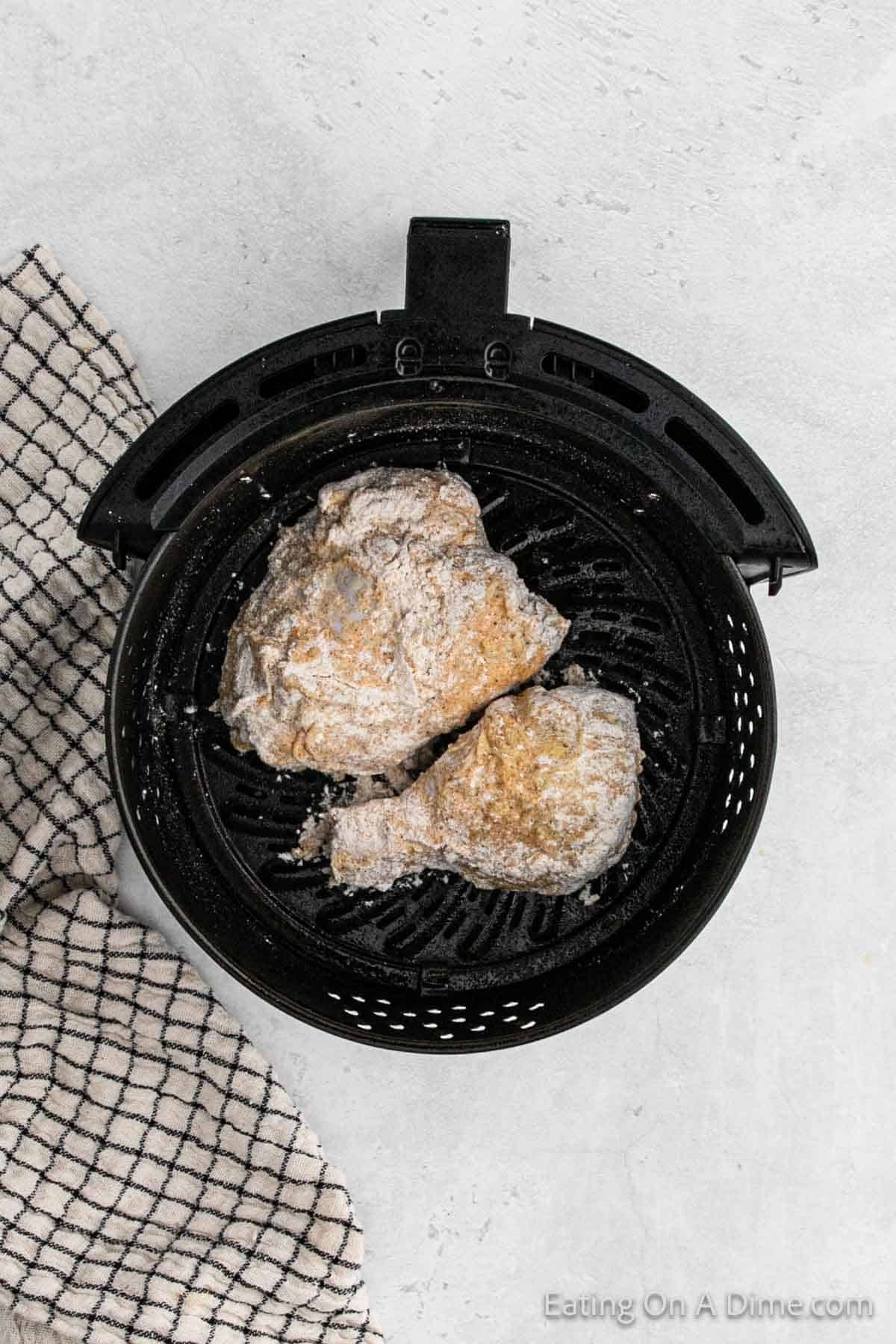 Two pieces of breaded chicken placed in an air fryer basket, ready for cooking into delicious air fryer fried chicken. A black and white checkered kitchen towel is partially visible to the side of the air fryer on a light gray surface.