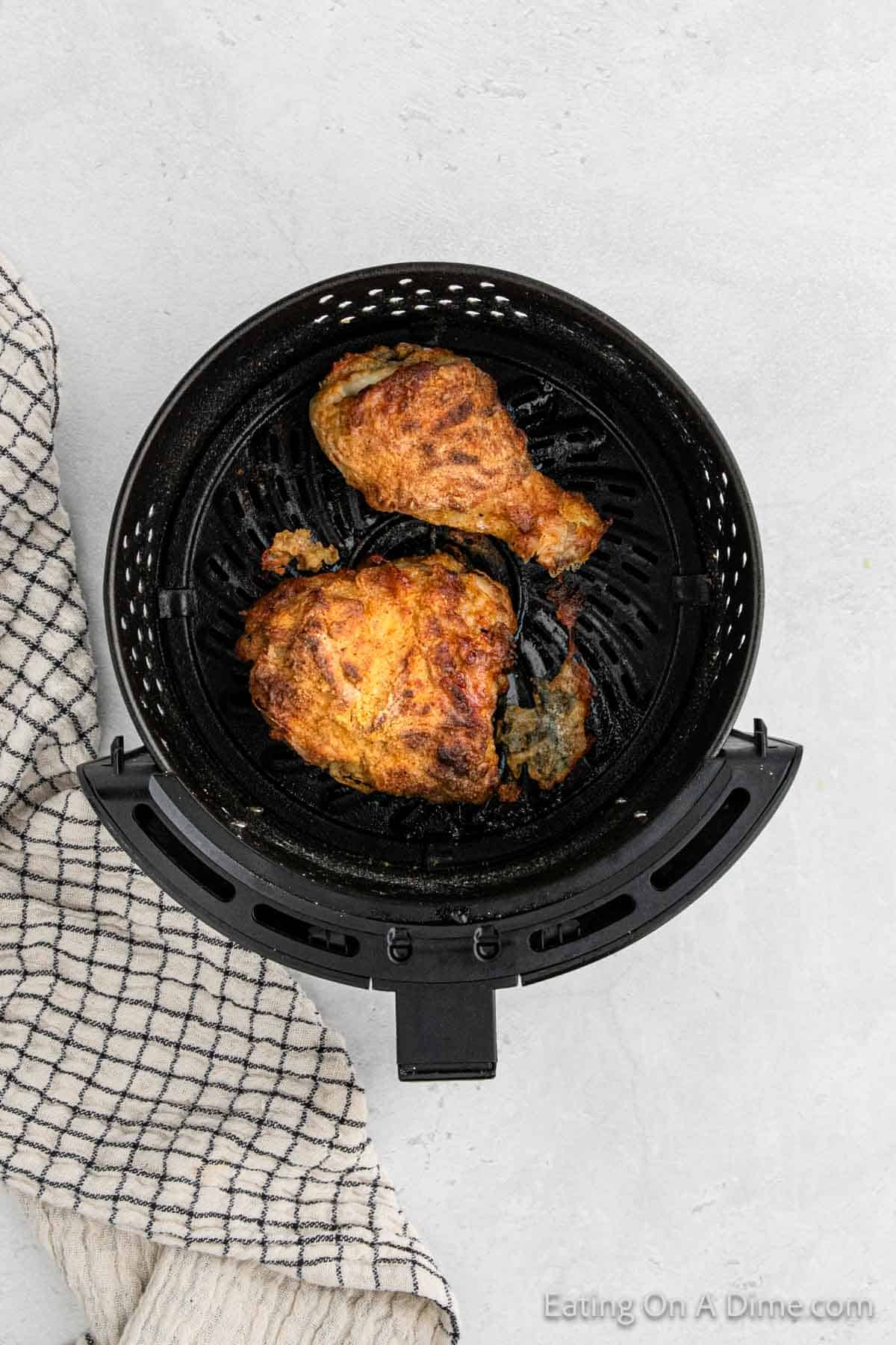 Overhead view of roasted chicken pieces inside an air fryer basket. The air fryer fried chicken appears well-cooked with a golden-brown, crispy finish. A checkered dish towel is partially visible on the left side, set on a light gray surface.