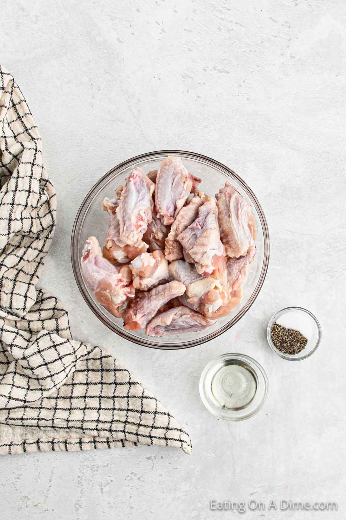 Uncooked chicken wings in a bowl with small bowls of oil and salt and pepper
