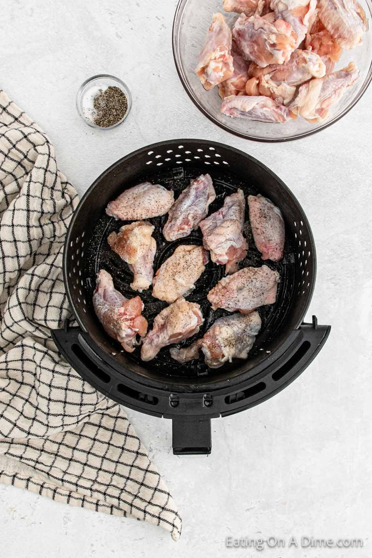 Placing chicken wings in the air fryer basket with a bowl of uncooked chicken wings on the side and a bowl of salt and pepper
