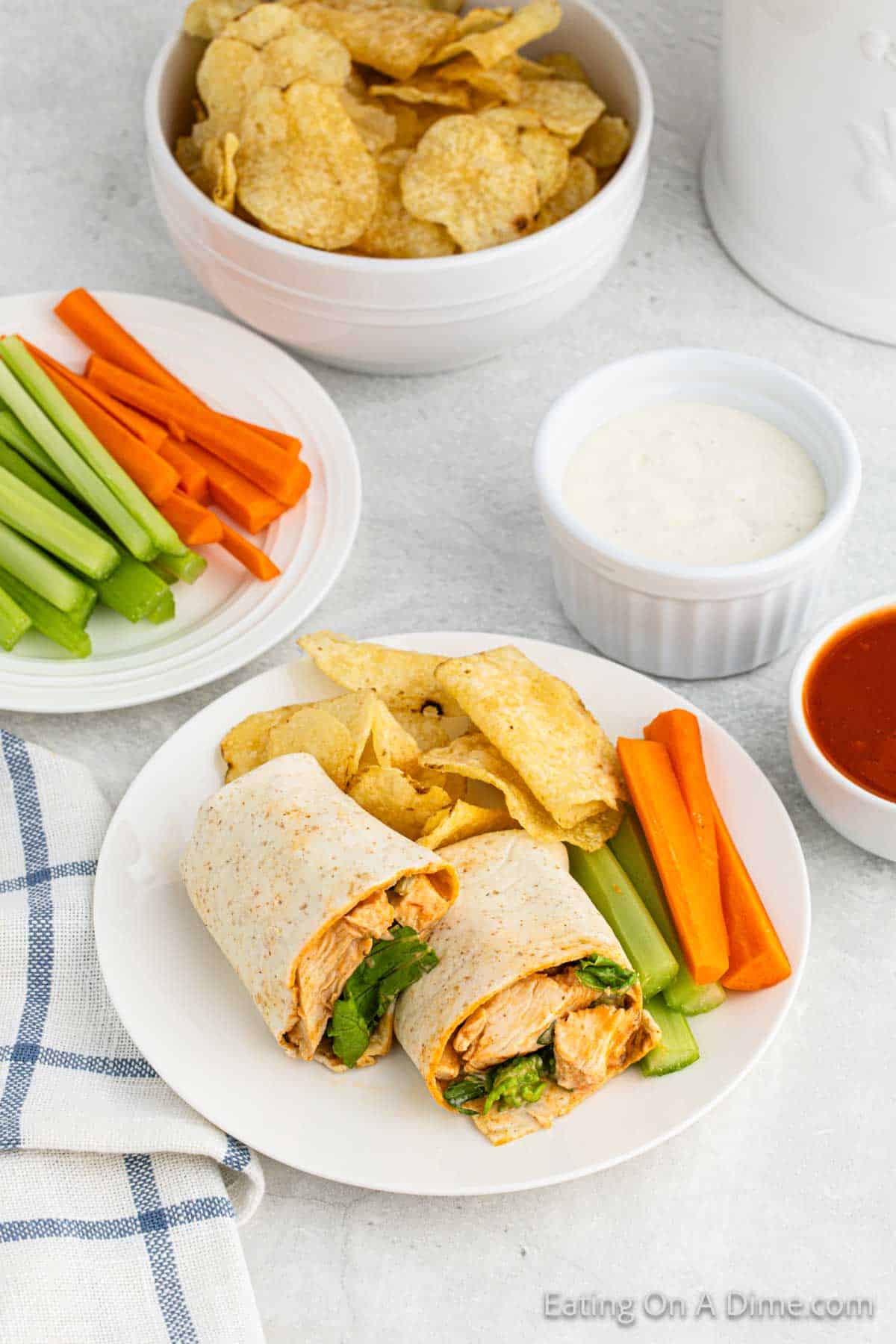 Buffalo Chicken wraps cut in half with a side of potato chips and celery and carrots with a plate of celery and carrots with a bowl of ranch dressing and buffalo sauce