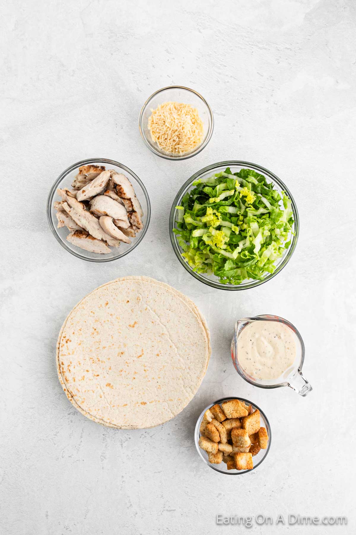Ingredients - Tortilla on a plate, grilled chicken strips, shredded lettuce, caesar salad dressing, croutons and parmesan cheese