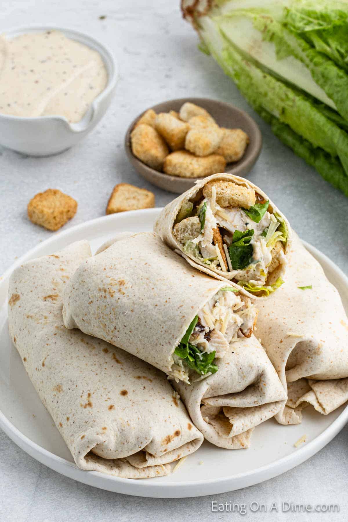 Chicken Caesar Wrap cut in half stacked on a plate with a bowl of croutons on the side