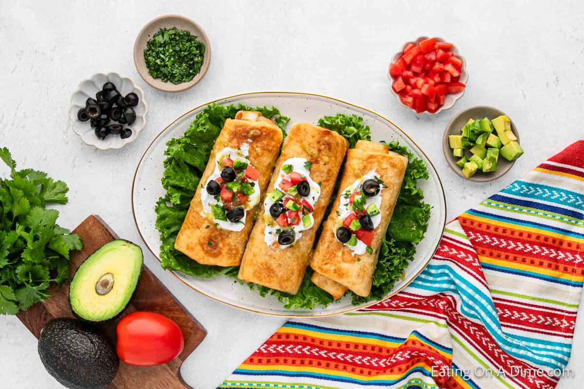 3 Crispy chimichangas on lettuce on a platter with bowls of diced tomatoes, diced avocado, slice black olives 