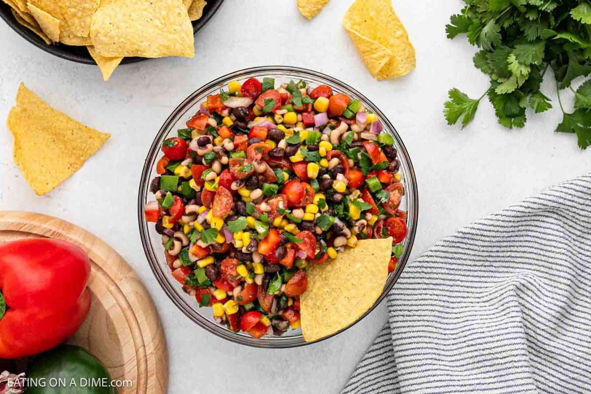 A bowl of Texas caviar sits on a table, bursting with black-eyed peas, corn, diced tomatoes, and onions. A tortilla chip is dipped into this colorful salsa. Surrounding the bowl are more tortilla chips, a red bell pepper, and a sprig of cilantro.