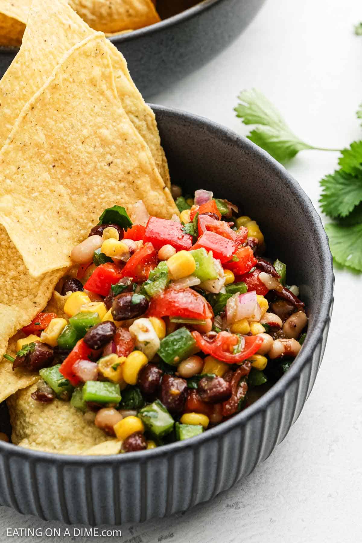 A bowl filled with vibrant Texas caviar, featuring black beans, corn, tomatoes, and green peppers, is served with tortilla chips. Fresh cilantro is visible beside the bowl on a white surface.