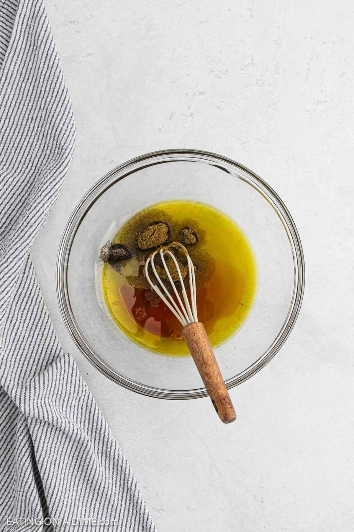 A glass bowl containing olive oil, vinegar, and spices sits with a whisk on a light gray textured surface, perfect for preparing Texas caviar. A gray and white striped cloth is draped to the left of the bowl.