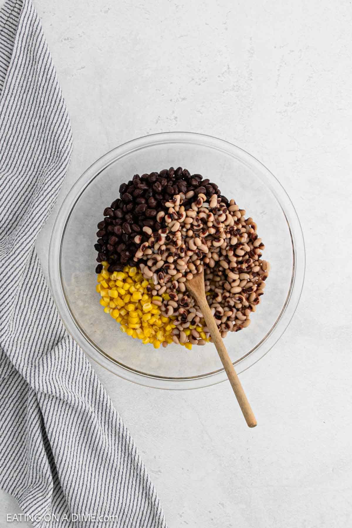 A glass bowl brimming with Texas caviar—black beans, black-eyed peas, and corn—sits on a light surface. A wooden spoon rests inside the bowl, while a striped kitchen towel is laid nearby.