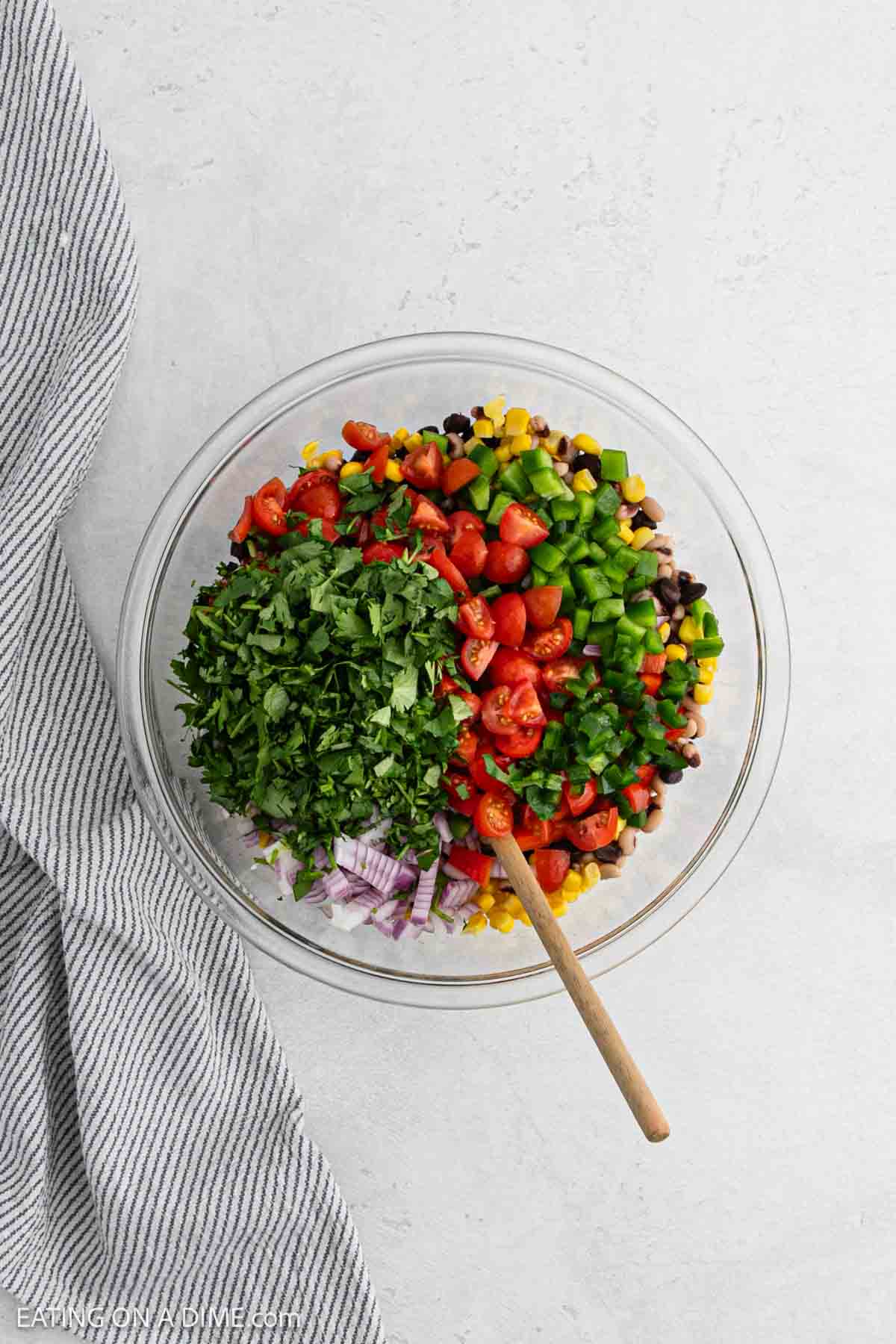 A glass bowl brims with vibrant Texas caviar, featuring cherry tomatoes, green bell peppers, black beans, corn, red onion, and cilantro. A wooden spoon rests inside the bowl. A striped cloth is placed beside it on a light background.