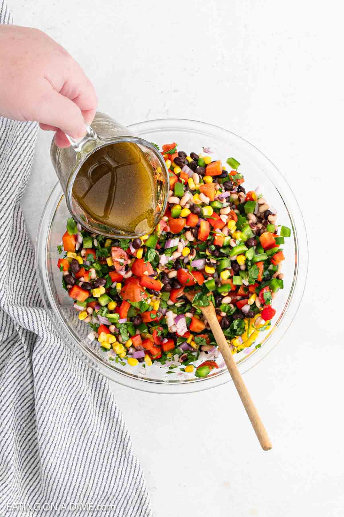 A hand pours dressing from a glass pitcher over Texas caviar in a clear bowl. This vibrant salad features corn, black beans, tomatoes, red onion, and chopped greens. A wooden spoon rests in the bowl, with a striped cloth beside it on the pristine white surface.