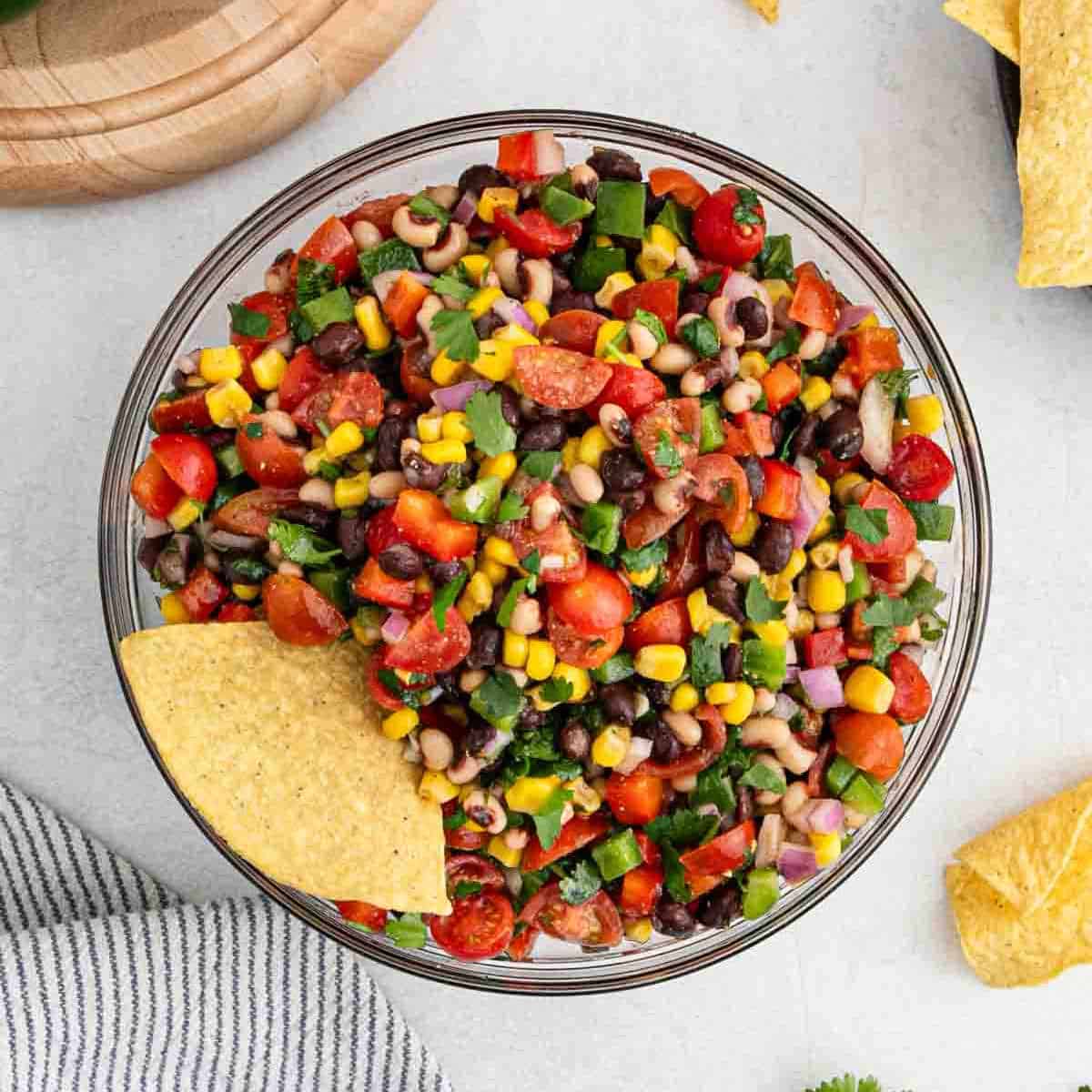 A glass bowl brimming with Texas caviar—a vibrant salad showcasing black-eyed peas, black beans, corn, diced tomatoes, bell peppers, and onions. A tortilla chip is dipped into the mix. Nearby, a wooden cutting board and striped cloth add a rustic touch.