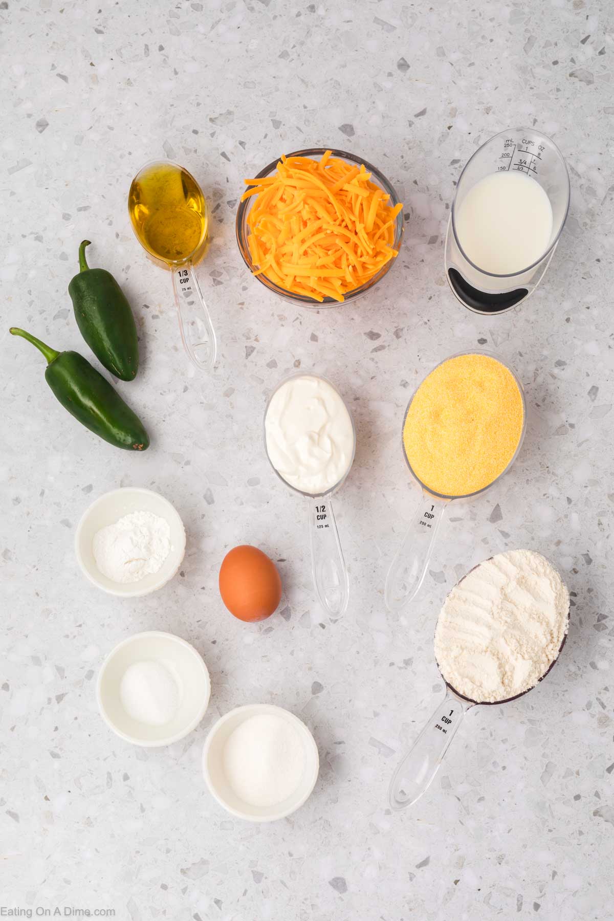 Ingredients arranged on a countertop for making jalapeno cheddar cornbread. Items include two whole jalapeños, shredded cheddar, milk, oil, sour cream, cornmeal, flour, sugar, baking powder, salt, and an egg. Measuring cups and spoons are used for some ingredients.