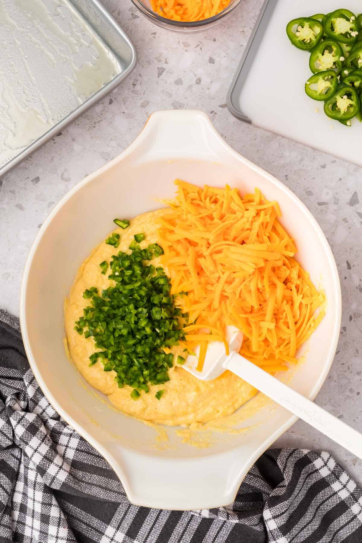 A white mixing bowl filled with jalapeno cheddar cornbread batter, shredded cheddar cheese, and chopped jalapeños rests on a gray countertop. A white spatula is in the bowl. Nearby are sliced jalapeños, shredded cheese, a baking dish, and a black-and-white checkered cloth.