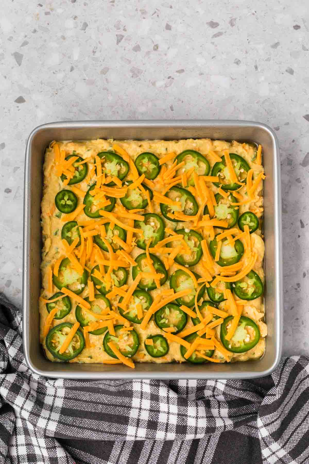 A square baking dish filled with jalapeno cheddar cornbread batter, topped with sliced jalapeños and shredded cheddar cheese. The dish is placed on a terrazzo countertop with a black-and-white checkered cloth draped nearby.