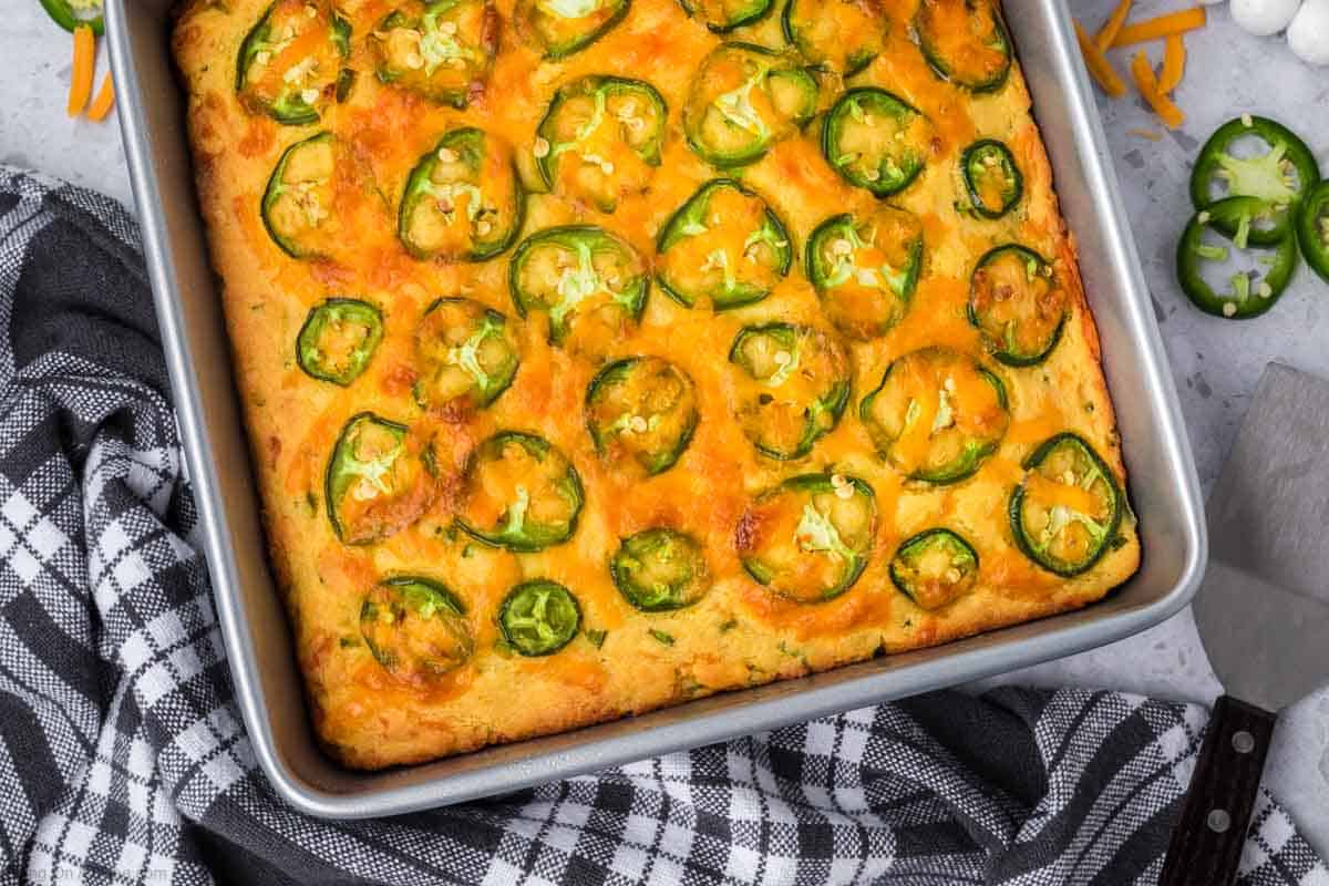 A square baking dish filled with golden-brown jalapeno cheddar cornbread, topped with sliced jalapeños. The dish is placed on a black and white checkered kitchen towel, and there is a metal spatula alongside it. Shredded cheddar cheese and additional jalapeño slices are visible nearby.