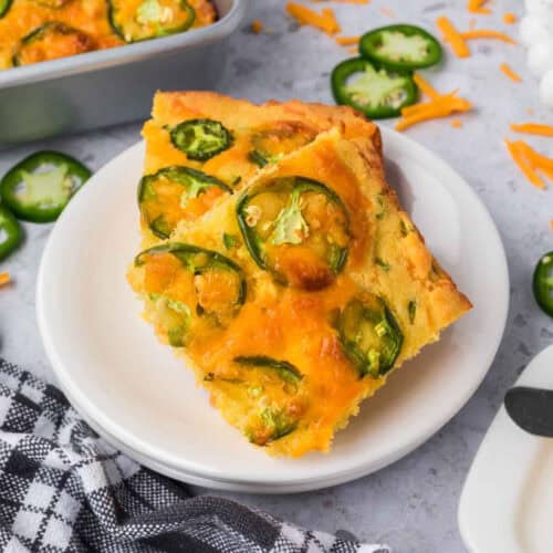 Close-up of two slices of jalapeno cheddar cornbread on a white plate. The cornbread is topped with jalapeño slices and melted cheddar cheese. A black and white checkered cloth, a baking pan with more cornbread, and scattered jalapeño slices and cheese are in the background.