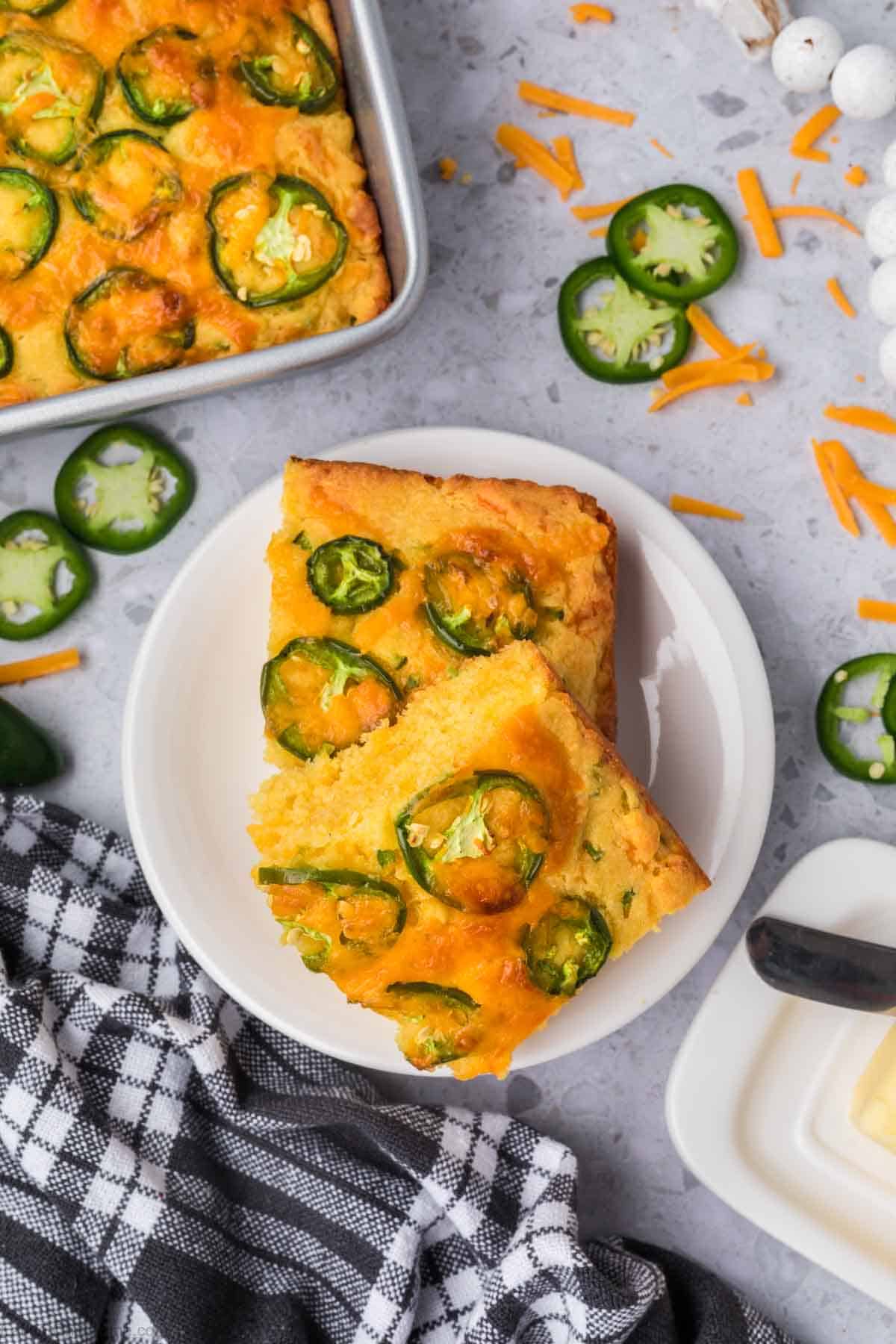 A white plate holds two pieces of jalapeno cheddar cornbread topped with melted cheese. The plate is on a gray countertop with scattered jalapeño slices, shredded cheese, and a black-and-white checkered napkin. A pan of cornbread and a butter dish are nearby.