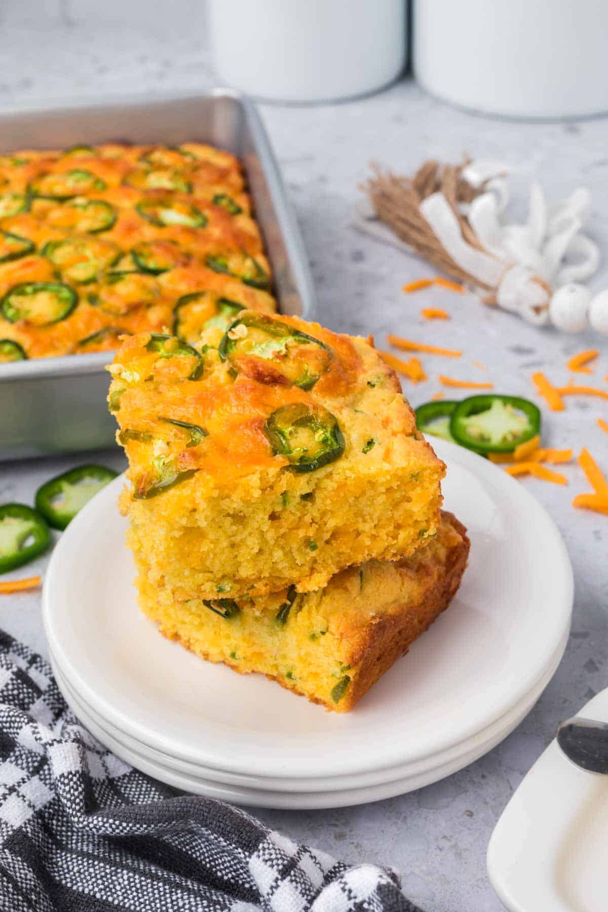 Two pieces of jalapeño cheddar cornbread stacked on a white plate, topped with slices of fresh jalapeño. In the background, a baking pan with more cornbread is visible, along with grated cheese, a black and white checkered cloth, and small decorative garlic bulbs.