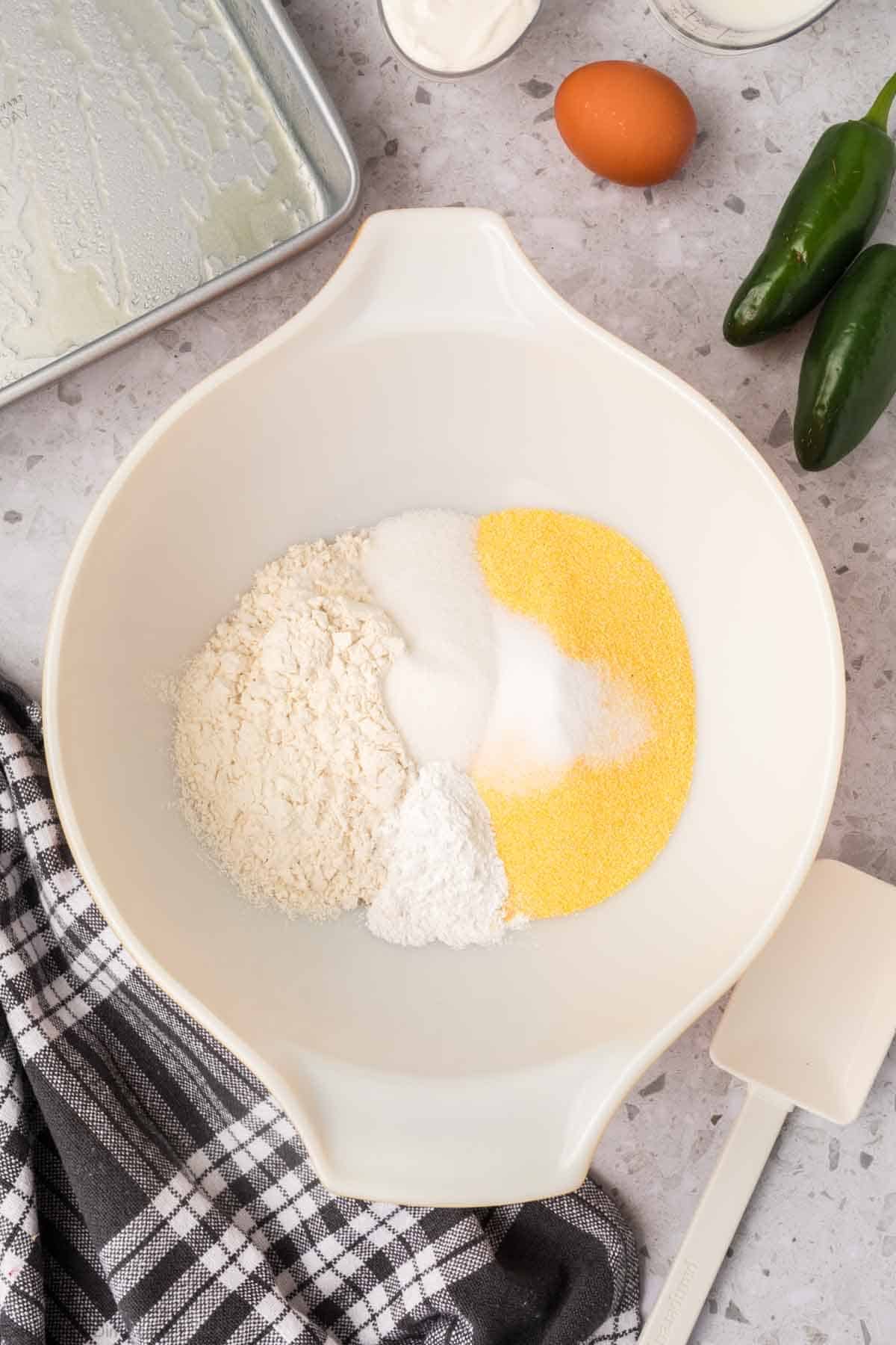 A white mixing bowl contains separate heaps of flour, cornmeal, sugar, and baking powder. A white spatula and a black and white checkered cloth are nearby. An egg, jalapeños, and cheddar sit ready for jalapeno cheddar cornbread in a greased baking pan positioned around the bowl on a speckled countertop.