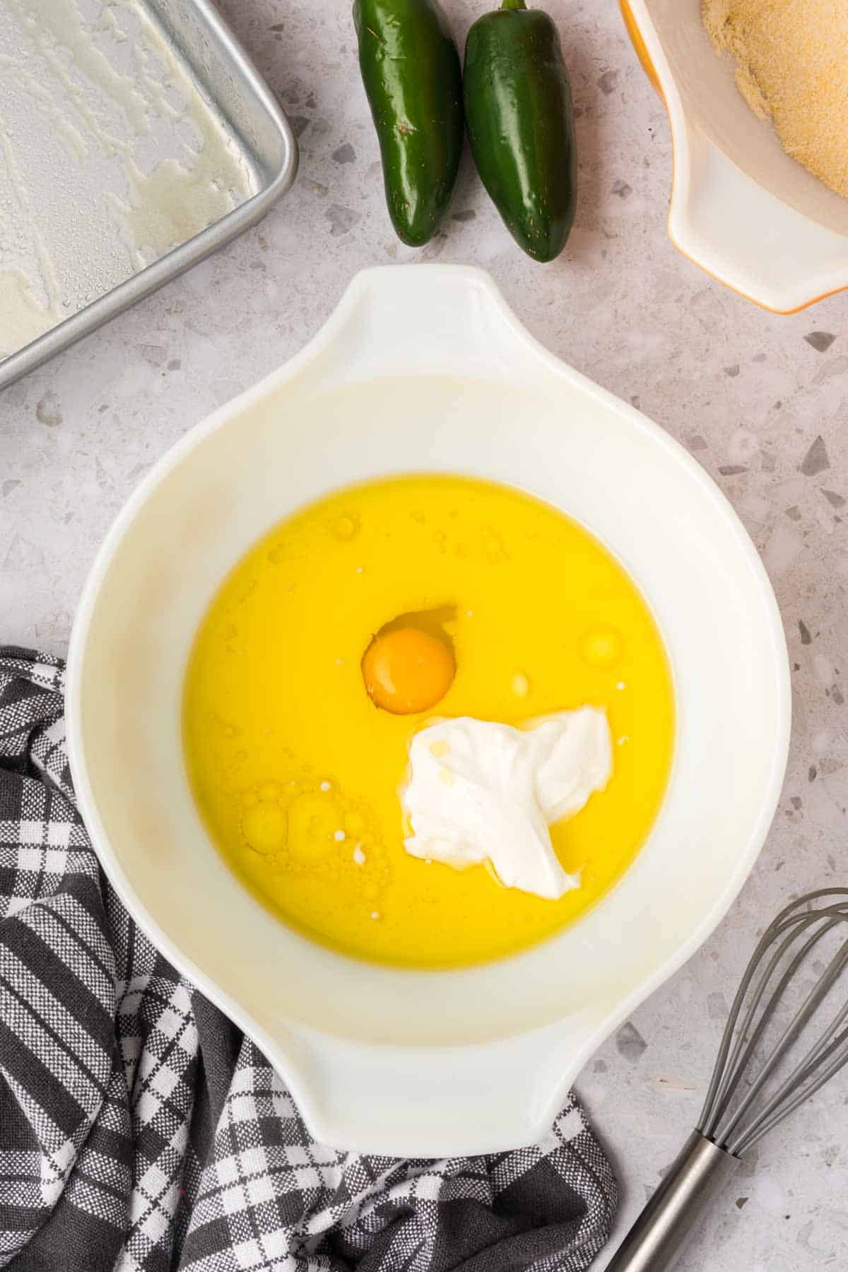 A large white mixing bowl contains a raw egg, melted butter, and a dollop of sour cream. Nearby, there are two jalapeños, a baking dish, a whisk, and a kitchen towel with a black and white checkered pattern—all the essentials for baking delicious jalapeno cheddar cornbread.