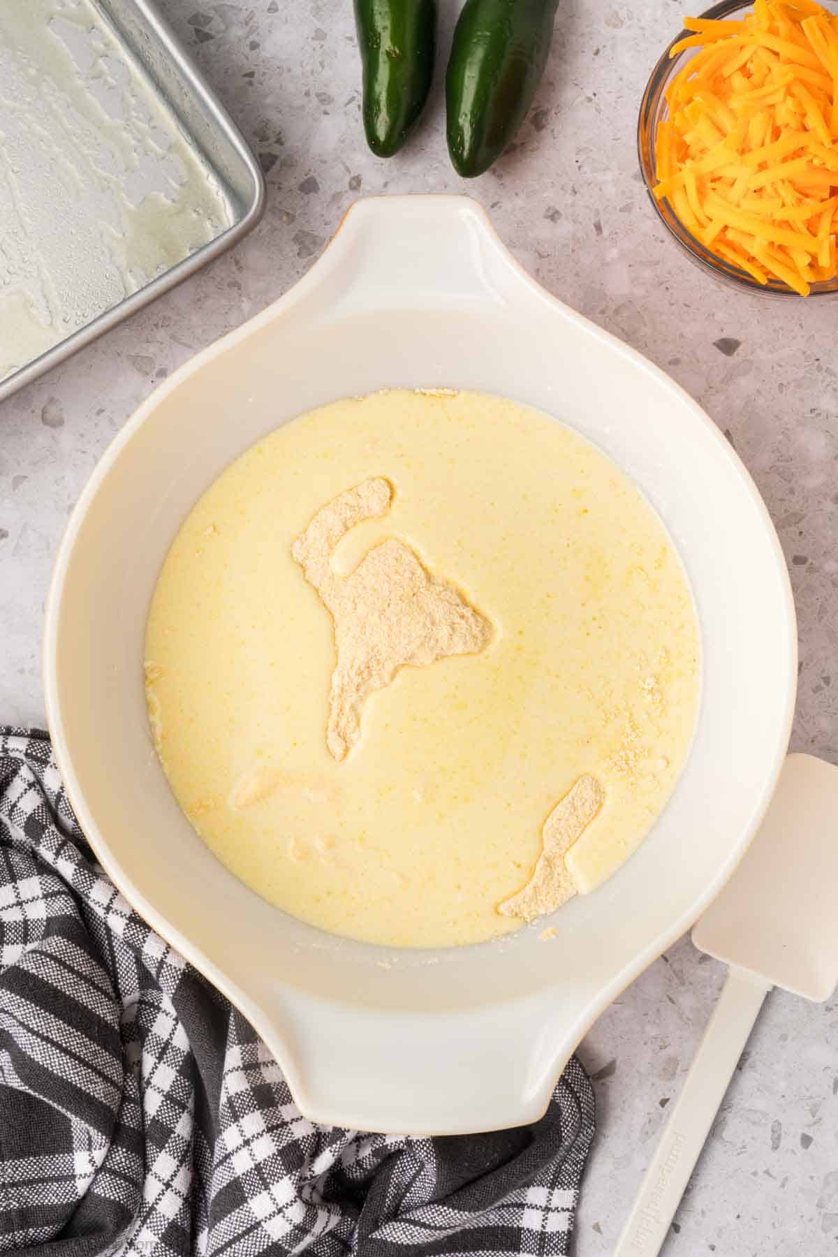 A mixing bowl filled with jalapeno cheddar cornbread batter sits on a gray countertop. A gray and white checkered cloth and a white spatula are next to the bowl. Shredded cheddar cheese and two green jalapeños are in the background near a lightly greased baking pan.