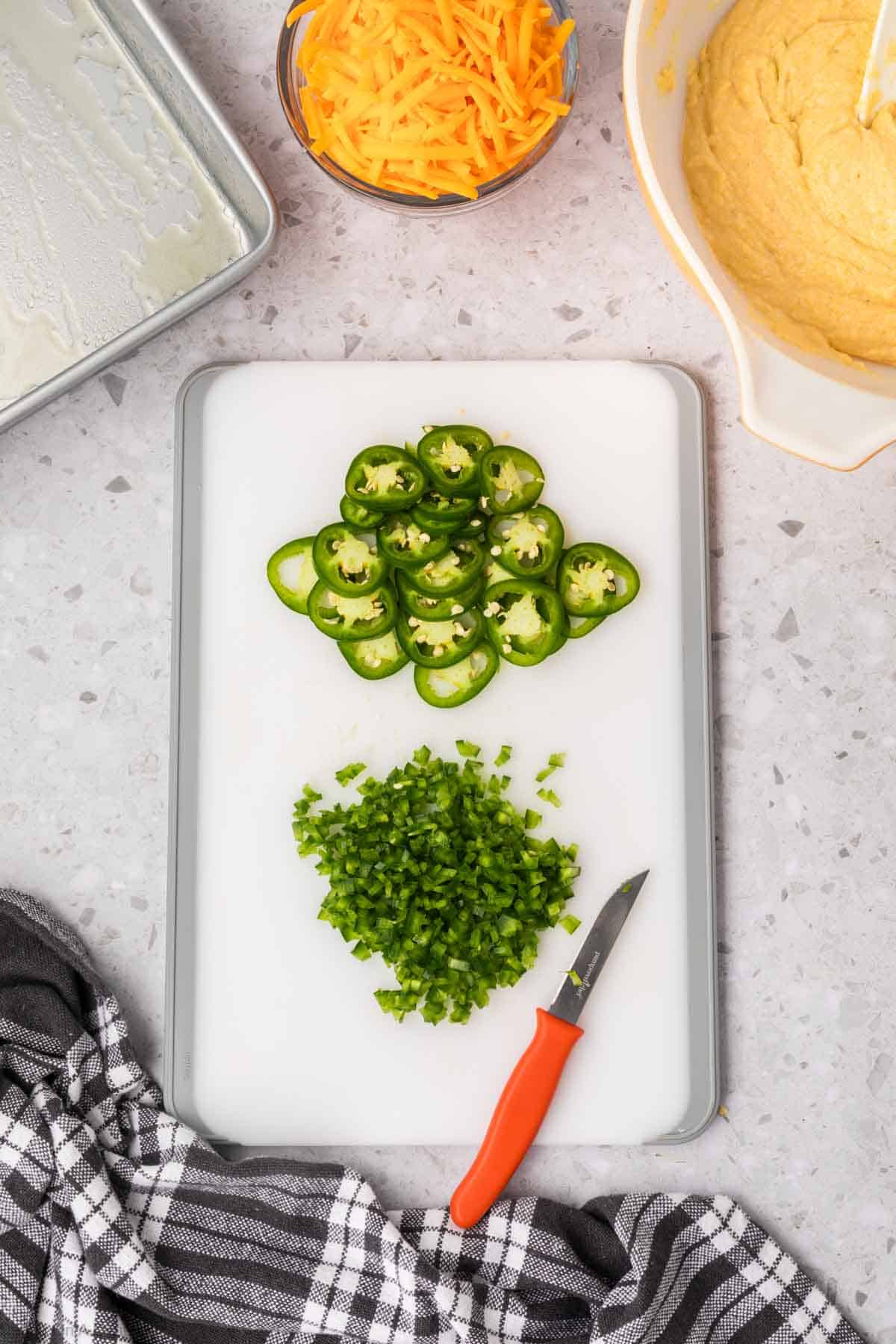 A cutting board with sliced and diced green jalapeños next to a knife with a red handle. Nearby, there is a bowl of grated cheddar cheese, a bowl of cornbread batter, and a greased baking pan—everything you need for jalapeno cheddar cornbread. A black and white checkered cloth is at the bottom left corner.