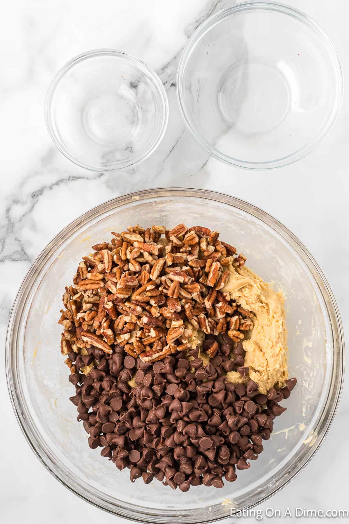 A clear glass bowl filled with cookie dough, chopped pecans, and chocolate chips sits on a marble countertop. Two smaller empty glass bowls are placed beside it. The image is from "Eating On A Dime.com," perfect for those looking to elevate their Chocolate Chip Cookie Cake recipe.