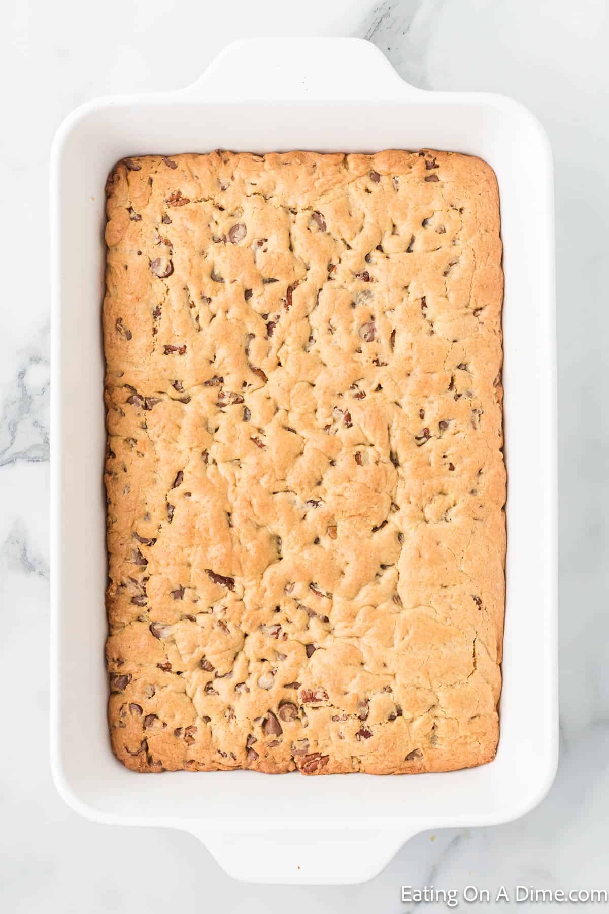 A rectangular white baking dish with baked cookie bars, featuring a golden-brown surface speckled with melted chocolate chips, embodies the quintessential Chocolate Chip Cookie Cake Recipe. The dish is placed on a light marble countertop.