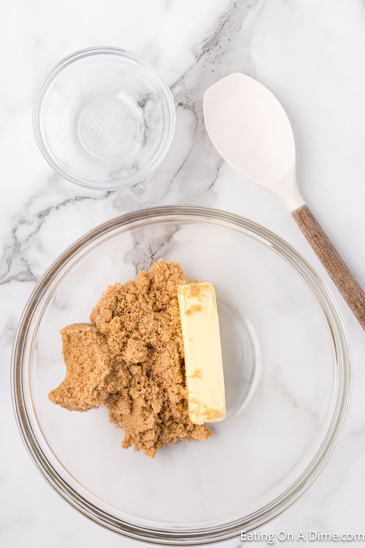 A clear glass bowl holds a stick of butter and brown sugar on a marble countertop, essential ingredients for a delightful Chocolate Chip Cookie Cake Recipe. Nearby, an empty smaller glass bowl and a white mixing spoon with a wooden handle await their turn to join the baking process.