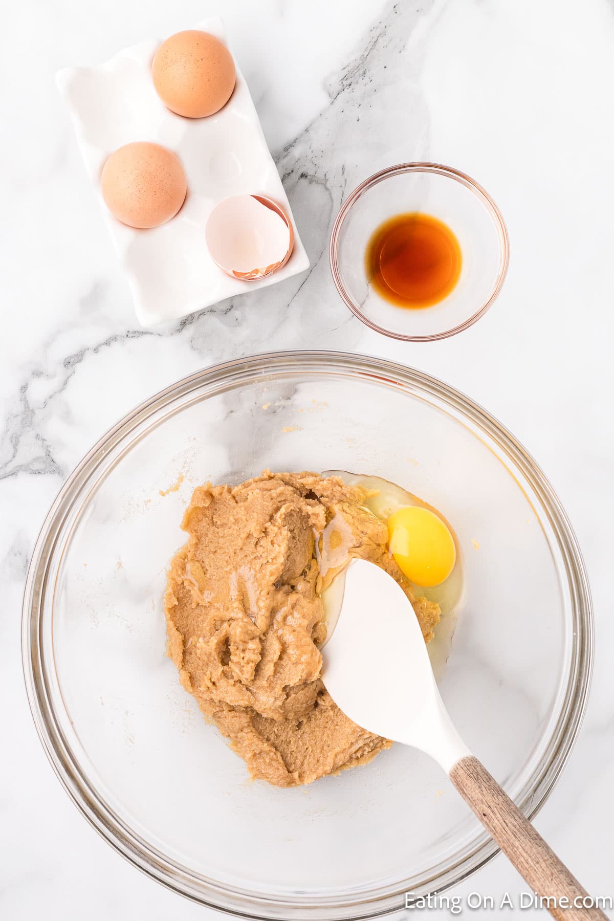 A marble countertop holds a mixing bowl with dough mixture, an egg, and a white spatula. To the side, there's a small bowl of vanilla extract and a ceramic dish with two whole brown eggs and one cracked shell—perfect for starting your Chocolate Chip Cookie Cake recipe.