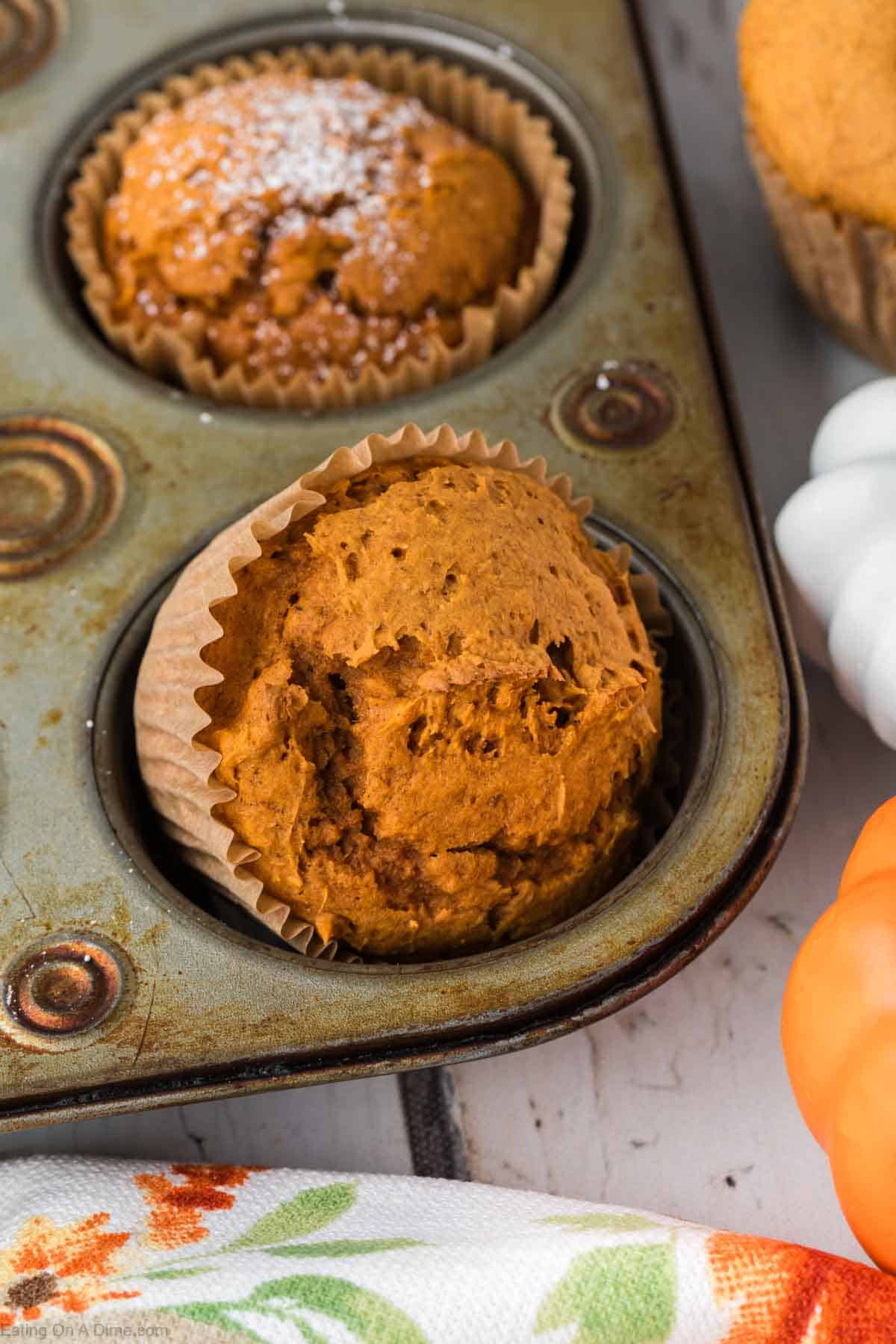 Close-up of a muffin pan with two 2 Ingredient Pumpkin Muffins, one sprinkled with powdered sugar and one plain, nestled in paper liners. The pan is slightly worn, adding rustic charm. A small white pumpkin and part of an orange towel with floral patterns are visible in the background.